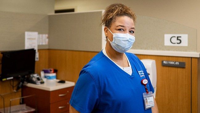 Phlebotomist Victoria Stephen stands in an office wearing blue scrubs and a blue face mask