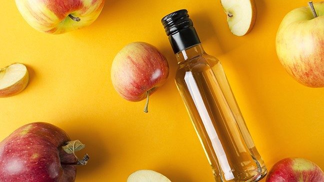 A glass container filled with apple cider vinegar sits atop a yellow surface surrounded by whole and sliced apples