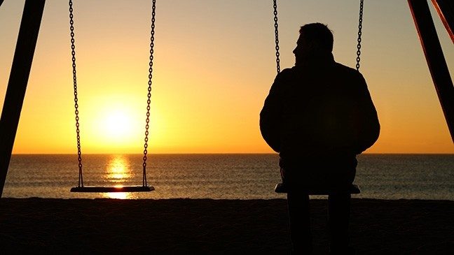 The silhouette of a man watching the sunset from a swing set with an empty swing next to him.
